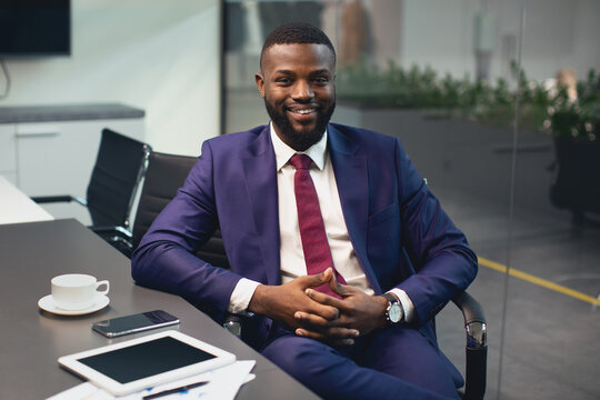 Handsome african american young businessman in nice suit sitting at table with documents, smartphone, digital tablet, drinking coffee at conference hall in office, having business meeting, copy space