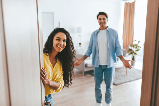 Two friends warmly greet each other in a bright and cozy living room. The woman smiles from behind the door while the man welcomes her with open arms, creating a joyful atmosphere.