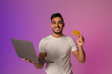 Millennial Arab man smiles while holding a credit card and laptop. He stands in neon light, showcasing a modern approach to remote banking and online shopping with contactless payments.