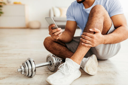 A man sits on the floor in casual clothing, checking his mobile phone after exercising with a dumbbell beside him. He appears focused and attentive to his fitness routine in a bright room. - Powered by Adobe