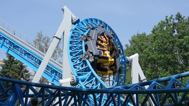 Roller coaster cart with people riding it goes through a loop upside down in green trees bushes city park. Loop metal frame of a roller coaster in an amusement park
