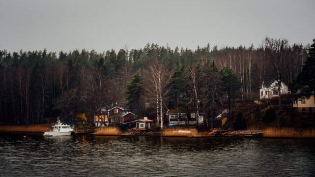 Scandinavian lakeside scenery with small wooden houses, forest and a boat near the shore on a calm overcast day. Moody Nordic landscape with quiet water, birch trees and cozy cabins in the distance.
