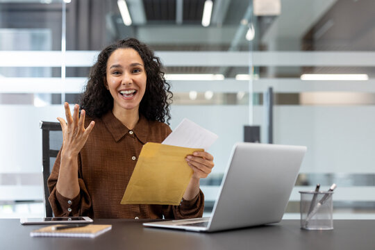 Happy businesswoman reading good news from a letter, celebrating promotion or success while working at her desk with a laptop in a modern office environment