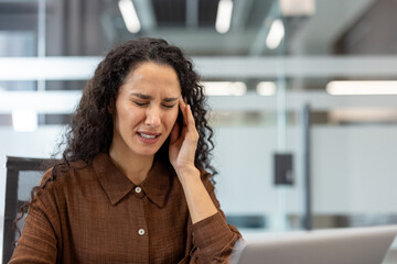 Young stressed businesswoman feeling intense pain and discomfort from a severe headache, pressing her temple while working on a laptop in a modern office environment