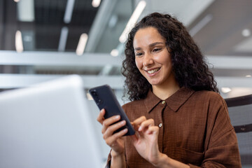 Young business woman with curly hair smiling while typing on her smartphone, connecting with people and managing tasks in a contemporary office environment
