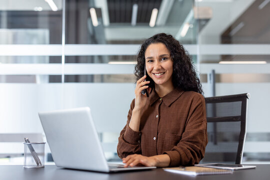 Young businesswoman communicating on a phone call, smiling while typing on her laptop at a modern office desk, demonstrating professionalism and productivity