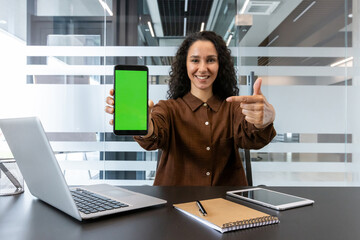 Businesswoman presenting new application on smartphone with green screen, sitting at office desk, smiling while showcasing mobile technology and digital content