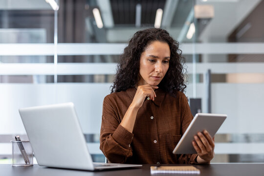 Professional businesswoman in smart casual attire reviewing data on a digital tablet at her desk with a laptop, focused and productive in a modern hybrid office workspace