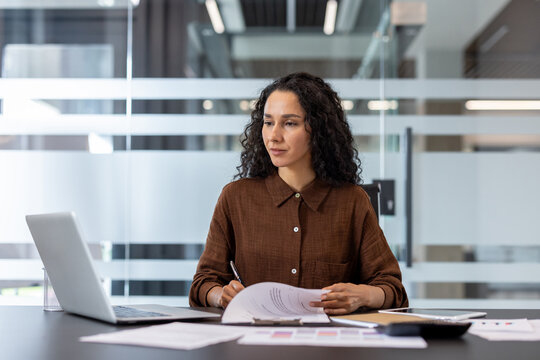 Young businesswoman with curly hair sitting at her office desk, looking focused while working with a laptop and analyzing paperwork and financial reports - Powered by Adobe