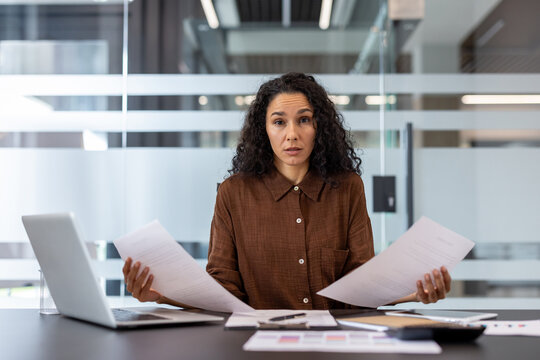 Stressed businesswoman sitting at a modern office desk, looking directly at the viewer with a concerned expression while holding papers, signifying workload, financial problems, and confusion