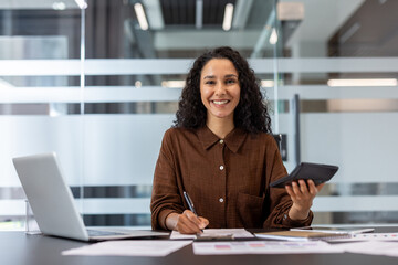 Young entrepreneur working on financial calculations and paperwork at office desk, holding a calculator and writing notes, exuding confidence and professionalism