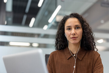 Woman with curly hair in brown shirt wearing earphones, looking at camera while using laptop for a focused online video conference in a modern office setting
