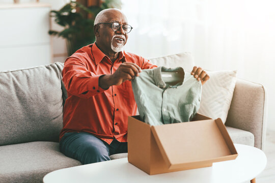 An elderly man sits on a couch with a joyful expression as he opens a package containing a green shirt. Sunlight streams through the window, highlighting the moment.
