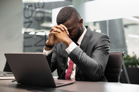 Upset african american man in suit CEO sitting with head down at workdesk in front of laptop at modern office, businessman suffering from financial crisis or bankruptcy, copy space