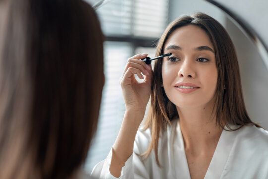 Beautiful millennial lady smiles at her reflection while applying mascara in her bathroom. Her daily makeup routine showcases her focus and care for her appearance, enhancing her features.