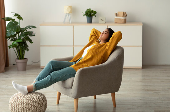 A relaxed Arab woman is sitting in a comfortable armchair with her hands behind her head and eyes closed. She enjoys her weekend, finding peace in her home environment.