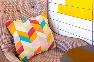Colorful geometric cushion on patterned armchair beside yellow and white tiled wall in modern living room or coffee shop, creating a modern and vibrant interior decor scene.