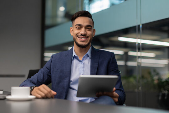 A cheerful middle eastern businessman in a neat suit sits at a table in a bright office. He is using a digital tablet to explore the latest business applications while smiling happily.