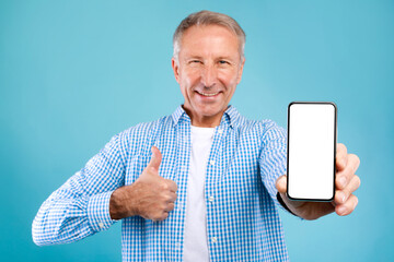 A cheerful senior man stands against a blue background, holding up a smartphone with a blank white screen. He gives a thumbs up, excited about a great mobile offer.