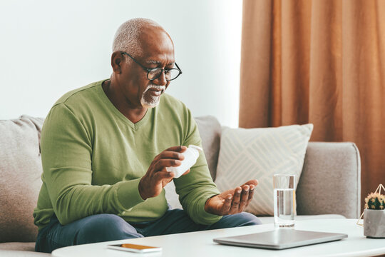 A senior man with glasses inspects his medication in a cozy living room. He is seated on a couch with a glass of water and a tablet nearby, reflecting a moment of health management.