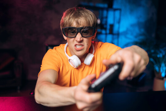 Focused, intense young male gamer in an orange shirt and protective glasses playing a console video game in a dark room with neon lights.