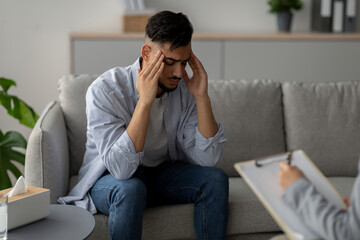 A stressed man sits on a couch, touching his temples in frustration. He is undergoing a therapy session with a psychologist, seeking help for his mental health challenges.