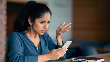 A woman appears frustrated while looking at her smartphone in a cafe. She seems confused as she gestures with her hand, surrounded by a calm atmosphere with books and glasses nearby.