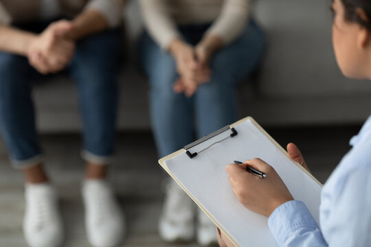 In a counseling session, a marital counselor takes notes on a clipboard while sitting across from a young couple. The atmosphere is calm, focusing on open communication and support.