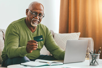 An older gentleman sits on a couch, holding a credit card while looking at a laptop. Natural light fills the room, creating a warm atmosphere. He appears focused on online shopping.