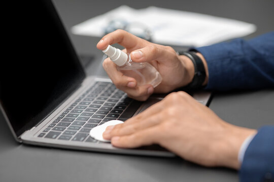 Unrecognizable man in a suit uses a cotton pad and alcohol gel to clean his laptop keyboard. This scene takes place in an office environment during the COVID-19 pandemic.