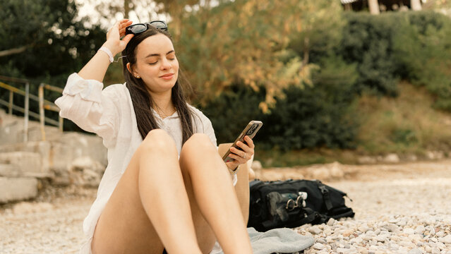 Woman sitting on a pebble beach, using her smartphone and enjoying a beautiful sunny day. Relaxing vacation moment by the sea, leisure and technology concept.