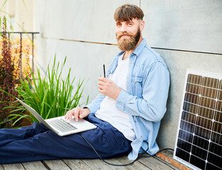 Portrait of barded man works on laptop on lap with glass of iced coffee. Solar panel nearby, emphasizing sustainable, modern eco-friendly workspace that combines technology and sustainability.