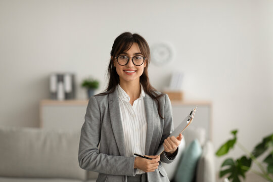 A joyful female psychologist poses with a clipboard in her modern office. She looks directly at the camera, exuding warmth and professionalism. Her workspace is inviting and thoughtfully designed. - Powered by Adobe