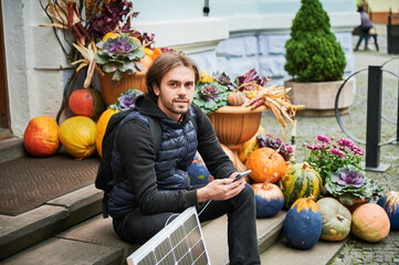 Handsome man using smartphone connected to photovoltaic solar panel. Integration of sustainable renewable energy into everyday life, demonstrating practical use of solar power for charging devices.