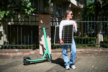 Woman using portable solar panel to charge scooter, highlighting integration of sustainable renewable energy. Green electric scooter connected to solar panel, emphasizing eco-friendly transportation.