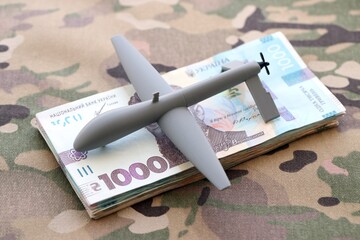Unnamed grey toy drone and ukrainian money bills on camouflage fabric close up. Drone manufacturing funds