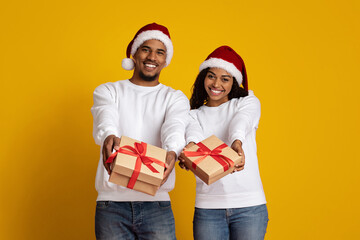 A joyful couple stands side by side, wearing matching Christmas hats. They each hold out a beautifully wrapped gift with festive ribbons, showcasing the spirit of holiday giving.