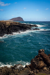Rocky volcanic coastline near Tenerife South Airport with strong waves and deep blue Atlantic water under a clear sky. Dramatic, wild and untouched coastal landscape.