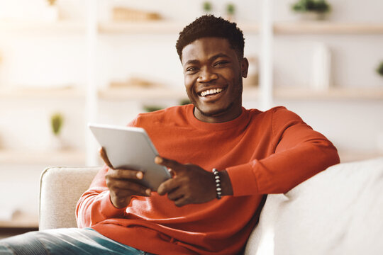 A cheerful young man sits comfortably on a couch, smiling while using a tablet in a bright and inviting living room filled with plants and soft light. - Powered by Adobe