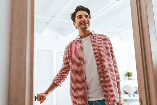 A man stands at the doorway, smiling happily. He is dressed casually in a checkered shirt and a plain t-shirt. The room features bright lighting and minimalistic decor.