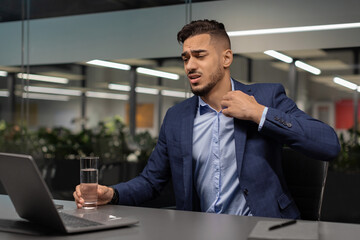 Fototapeta premium Young middle eastern businessman shows signs of distress while sitting at a worktable in a modern office. He is looking at his notebook screen and unbuttoning his shirt, holding a glass of water.