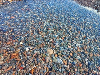 Clear shallow river water flowing over smooth multicolored stones, sunlight creating gentle reflections on the pebbles and natural rocky riverbed.