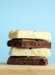 Stack of aerated white and milk chocolate bars on a wooden surface against a light blue background. The close-up highlights the bubbly texture and contrasting colors, creating a clean, minimal, and ap