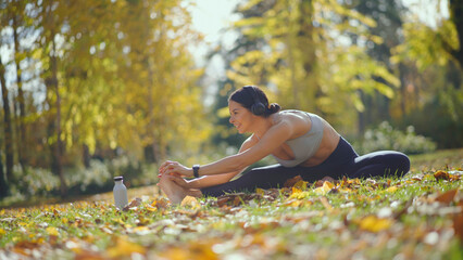 Woman engages in a focused stretching pose rooted on lush grass and fallen autumn leaves, wearing...