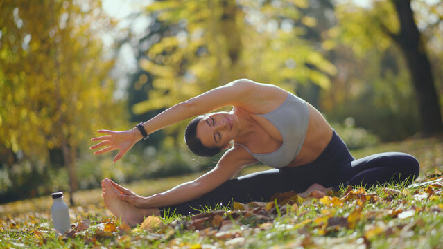 Woman practicing seated side-bend yoga on a mat in an autumn park, reaching overhead toward her feet among colorful fallen leaves, enjoying outdoor wellness and calm