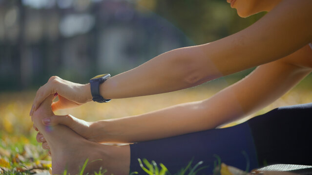 Woman performing a seated forward bend yoga pose in a park on a sunny day, focusing on flexibility and wellness for a healthy lifestyle, with a smartwatch tracking her fitness