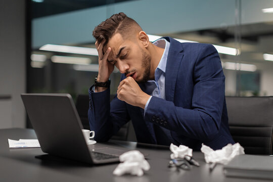 A young middle eastern businessman sits exhausted in his office, coughing and touching his head. He appears unwell, struggling with symptoms of coronavirus while working at his computer.
