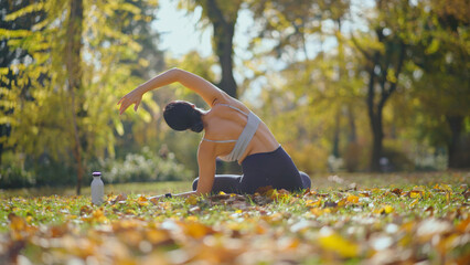 Woman performing a yoga pose, stretching her back and arms outdoors on fallen autumn leaves in a green park, focusing on mindfulness, exercise, and well-being