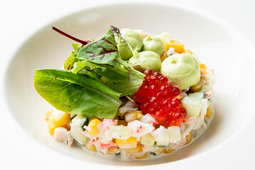 A gourmet layered crab salad appetizer with corn, cucumber, and avocado cream, elegantly topped with bright red caviar and fresh greens in a white bowl