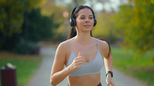 Woman jogging on a path in a park, wearing a sports bra and wireless headphones, with a smartwatch on her wrist while engaging in outdoor exercise and a healthy lifestyle